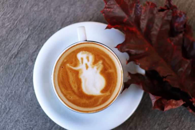Halloween B&B - white ceramic teacup filled with ghost illustration coffee latte on white ceramic saucer beside maroon leaf photography