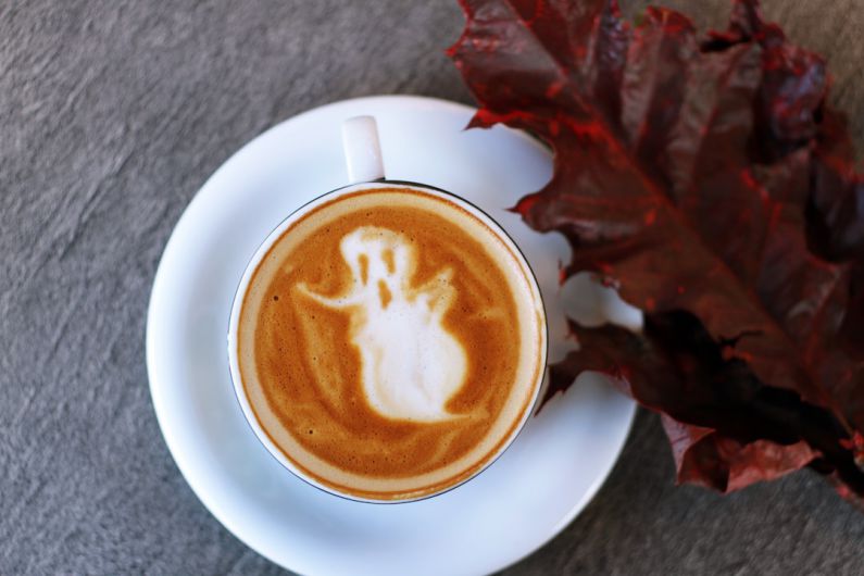 Halloween B&B - white ceramic teacup filled with ghost illustration coffee latte on white ceramic saucer beside maroon leaf photography
